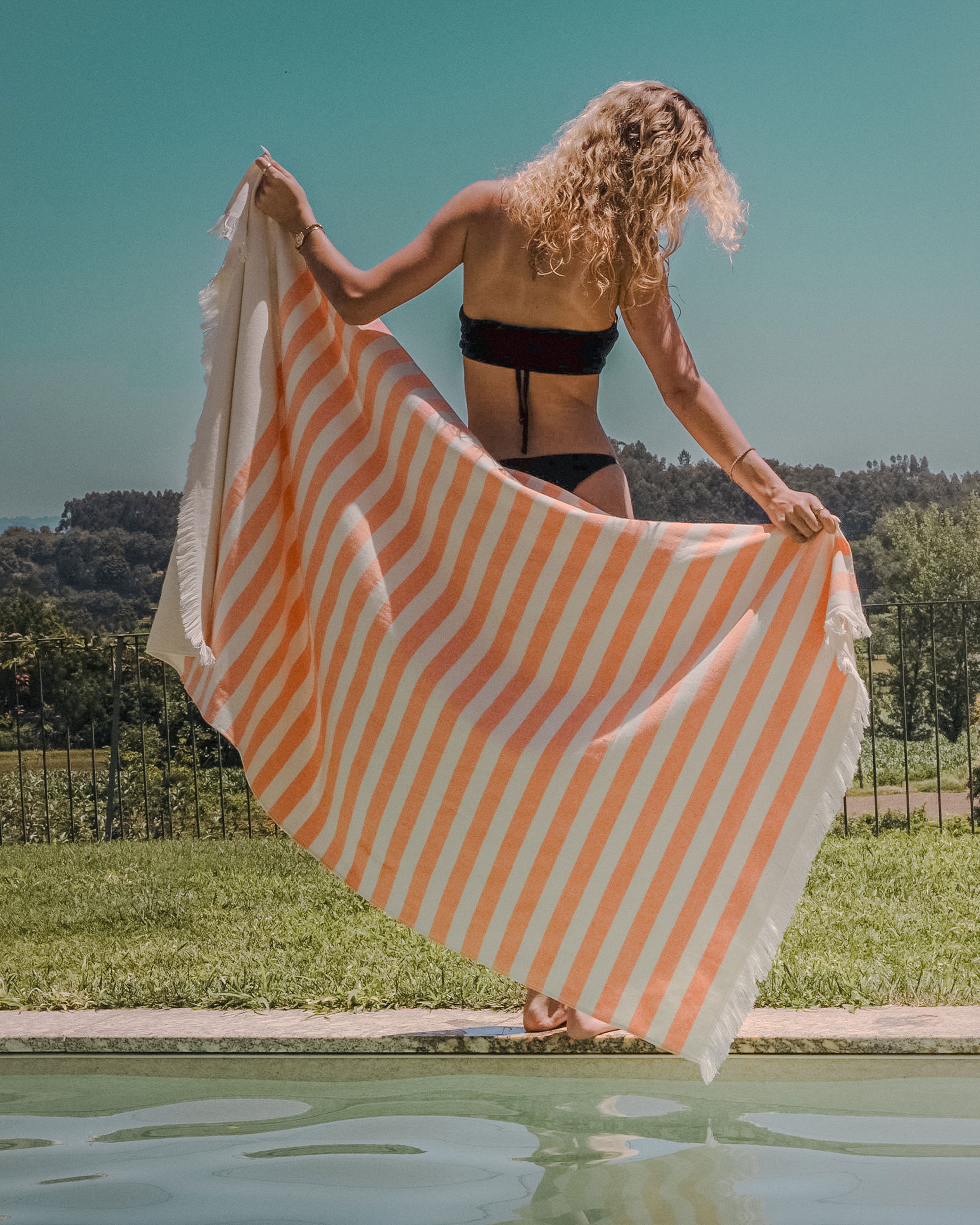 Woman holding an orange and white striped pareo by a pool with a scenic background
