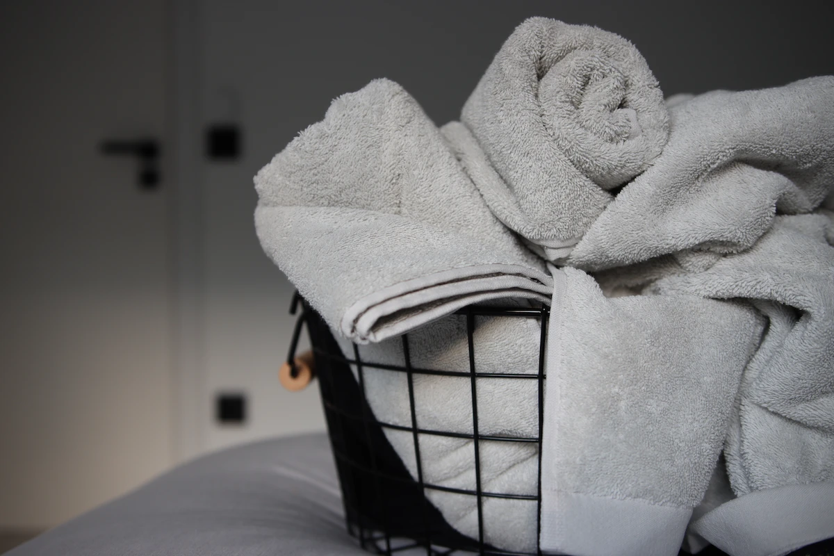 Schwarzer Drahtkorb, gefüllt mit grauen gewaschenen Handtüchern von Domsoeiro, auf einem Bett mit grauen Bettlaken, um sie weich zu halten. In der Zwischenzeit befindet sich im Hintergrund eine weiße Tür mit einem schwarzen Griff. Schwarze Steckdose und ein schwarzer Schalter an der weißen Wand.